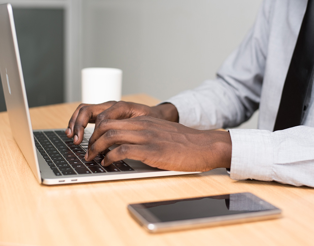 A man types on his laptop at a wooden desk.