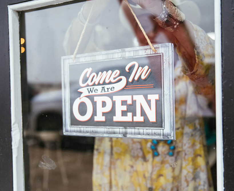 A woman hangs a "Come In, We're Open" sign in a storefront window.