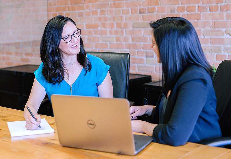 Two women meet at a table to look over papers and documents on a laptop.