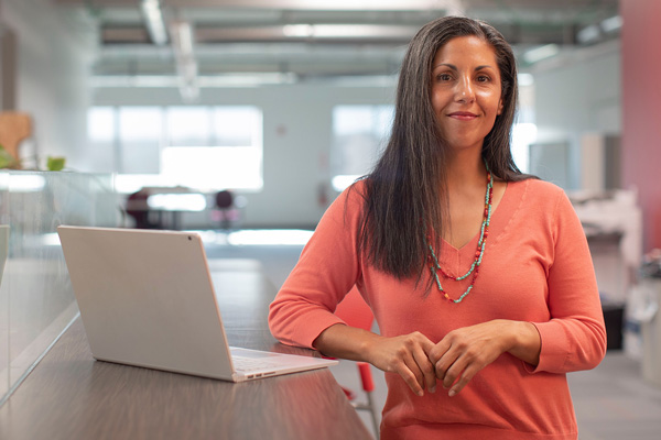 A woman stands by a desk with a laptop in an office environment.